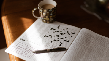 Magazine with crossword laid out on table with coffee mug and pen on top of it.